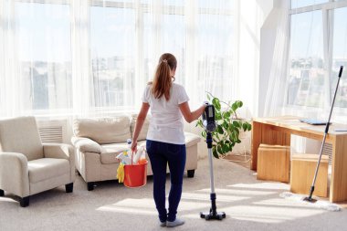 Beyaz gömlekli ve elinde kablosuz elektrikli süpürge ve deterjan tutan kot pantolonlu genç bir kadının arka plan görüntüsü. Ev işleri, temizlik ve ev işleri konsepti