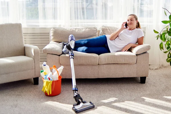 白いシャツを着た若い女性がソファに横たわって 自宅で掃除しながら携帯電話で話し スペースをコピーします 家事や家事の概念 掃除機 — ストック写真
