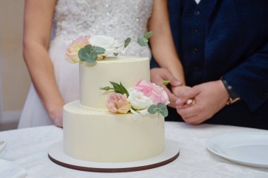 Restorandaki masada pembe ve beyaz gül çiçekleriyle süslenmiş zarif beyaz düğün pastası. Gelin ve damat arka planda, kopyalama alanı.