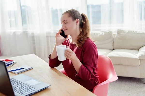 木製の机に座ってお茶を楽しみ 自宅のオフィスで携帯電話で話す幸せな若いビジネス女性 コピースペース — ストック写真