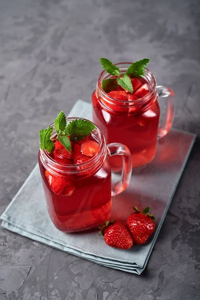 Homemade strawberry tea with mint in mason jar on gray concrete table background, copy space. Cold summer berry drink in sparkling glasses. Fresh vitamin beverage