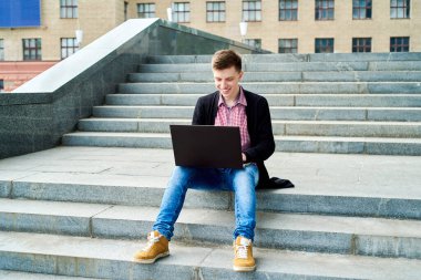 Üniversite kampüsünde oturan mutlu gülümseyen öğrenci dışarıda dizüstü bilgisayarla çalışıyor, kopyalama alanı. Sınava hazırlanmak, daktilo etmek, internette sörf yapmak. Teknoloji, eğitim kavramı