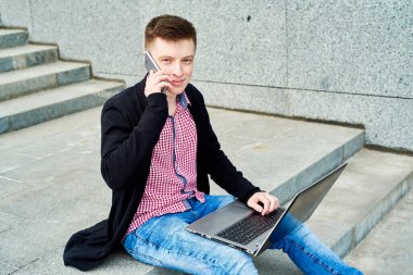 Mutlu gülümseyen öğrenci merdivenlerde oturuyor, dizüstü bilgisayarla çalışıyor ve cep telefonuyla dışarıda konuşuyor, kopyalama alanı. Sınava hazırlanmak, daktilo, internet. Teknoloji, eğitim kavramı