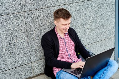 Üniversite kampüsünde oturan mutlu gülümseyen öğrenci dışarıda dizüstü bilgisayarla çalışıyor, kopyalama alanı. Sınava hazırlanmak, daktilo etmek, internette sörf yapmak. Teknoloji, eğitim kavramı