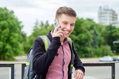 Sırt çantalı mutlu genç bir öğrencinin portresi. Şehirde yürüyor ve cep telefonuyla konuşuyor.