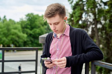 Parkta yürüyen, kulaklıkla müzik dinleyen ve açık havada cep telefonu kullanan mutlu genç bir öğrencinin portresi.