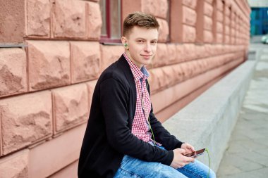 Taşın üzerinde oturan, kulaklıkla müzik dinleyen ve açık havada cep telefonu kullanan mutlu genç bir öğrencinin portresi.