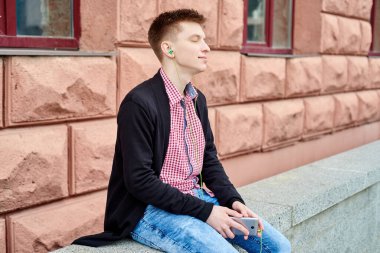 Taşın üzerinde oturan, kulaklıkla müzik dinleyen ve açık havada cep telefonu kullanan mutlu genç bir öğrencinin portresi.