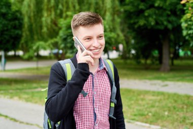 Sırt çantalı mutlu genç bir öğrencinin portresi parkta yürüyor ve cep telefonuyla dışarıda konuşuyor, boşluğu kopyalıyor.