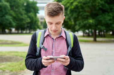Sırt çantalı mutlu genç bir öğrencinin portresi parkta yürüyor ve açık havada cep telefonu kullanıyor.