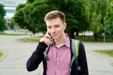 Sırt çantalı mutlu genç bir öğrencinin portresi parkta yürüyor ve cep telefonuyla konuşuyor, alanı kopyalıyor.