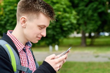 Sırt çantalı mutlu genç bir öğrencinin portresi parkta yürüyor ve açık havada cep telefonu kullanıyor.