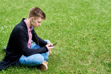 Parktaki yeşil çimlerde oturan, kulaklıkla müzik dinleyen ve dışarıda cep telefonu kullanan mutlu genç bir öğrencinin portresi.