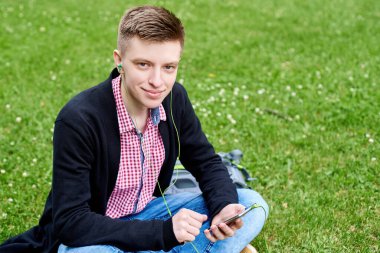 Parktaki yeşil çimlerde oturan, kulaklıkla müzik dinleyen ve dışarıda cep telefonu kullanan mutlu genç bir öğrencinin portresi.