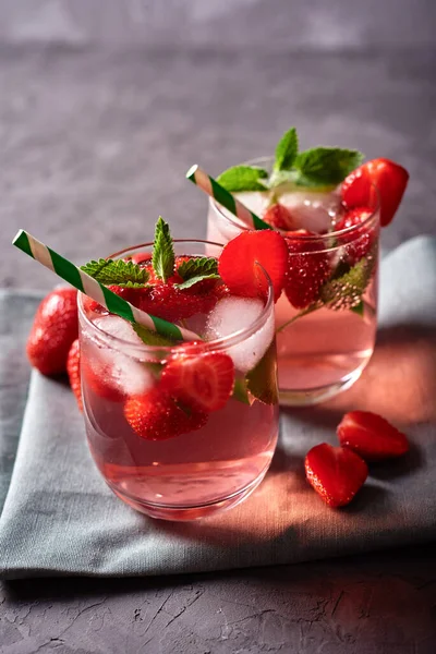 Fresh strawberry lemonade with ice, mint and paper straw in sparkling glasses on gray table background, copy space. Cold summer drink. Berry cocktail