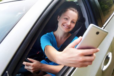 Cep telefonu tutan ve araba sürerken fotoğraf çeken mutlu genç bir kadın. Gülümseyen kız akıllı telefon kamerasıyla selfie çekiyor. Tatil ve turizm konsepti
