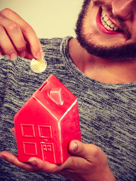 Smiling man putting coin into house piggybank