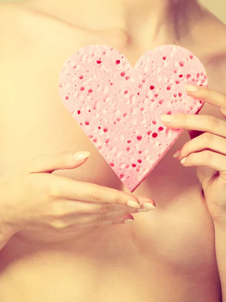 Naked woman holding pink heart sponge in hands.