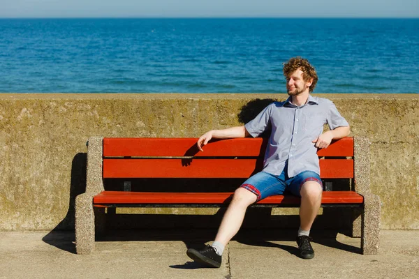 Szczęśliwy młody człowiek siedzący na ławce przez morze ocean. — Zdjęcie stockowe