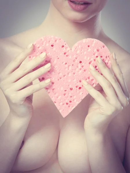 Naked woman holding pink heart sponge in hands.