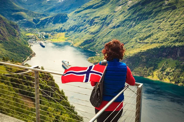 Female tourist with norwegian flag enjoying scenic view over fjord Geirangerfjord with cruise ship. Cruising vacation and travel.