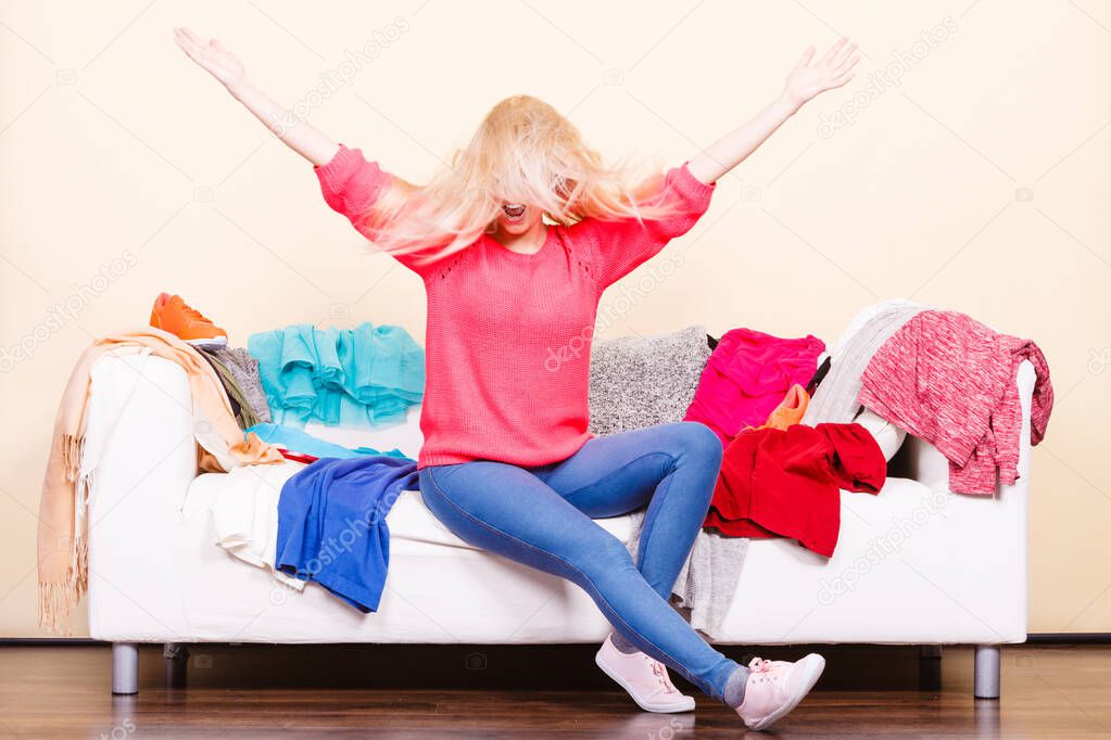 Clothing dilemmas concept. Blonde woman with windblown does not know what to wear sitting on messy couch with piles of clothes.
