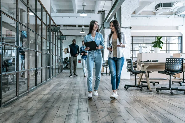 Condividere nuove notizie dall'ufficio. Due giovani colleghi in smart casual wear discutono di affari e sorridono mentre camminano per l'ufficio — Foto Stock