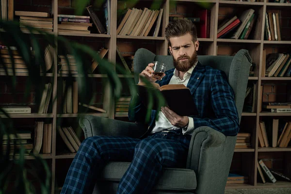 Elegante hombre en un traje con vaso de bebida sentado en habitación vintage y libro de lectura. Hombre de moda . — Foto de Stock