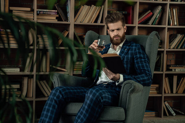 Elegant man in a suit with glass of beverage sitting in vintage room and reading book. Fashion man.
