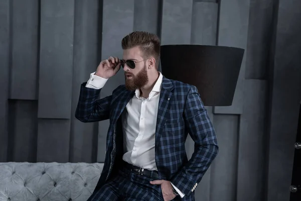 Hombre guapo y elegante en traje azul en una jaula y gafas en casa sentado en el sofá —  Fotos de Stock