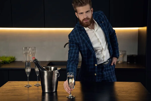 Hombre guapo y elegante en traje azul en jaula en casa. De pie en la moderna cocina negra con copa de champán — Foto de Stock