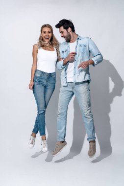 Gündelik beyaz tişörtler ve kotlar giyen, el ele tutuşup gri arka planda kameraya bakarken eğlenen, zıplayan ve dans eden neşeli modern çiftin yan görüntüsü..