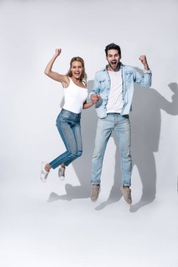 Gündelik beyaz tişörtler ve kotlar giyen, el ele tutuşup gri arka planda kameraya bakarken eğlenen, zıplayan ve dans eden neşeli modern çiftin yan görüntüsü..