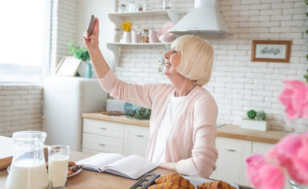 Attractive old woman cooking on kitchen. Cheerful grandmother baking and making selfie on a smart phone.