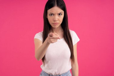 Korkmuş Asyalı kadın işaret parmağı ile kameraya doğru yatay görüntü, garip bir şey fark etmekten utanıyor, at kuyruğu ile taranmış koyu renk saçları var, pembe tişört giyiyor, pembe duvarda izole edilmiş..