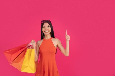 Güzel heyecanlı Asyalı kadın pembe duvar arkasında yalnız poz veriyor. Elinde alışveriş torbaları var. Logo için bedava spa. Turuncu elbiseli ve kafasında güneş gözlüğü olan..
