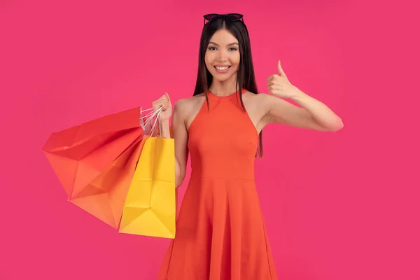 Beautiful happy asian woman posing isolated over pink wall background holding shopping bags pointing up, free spase for text ot logo. dressed in orange dress and sunglasses on head