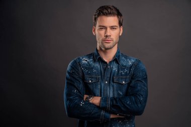 Yakışıklı genç adam kollarını kavuşturup kameraya bakarken gri arka planda duruyor..