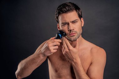 Yakışıklı, sakallı adam izole edilmiş. Üstsüz kaslı adamın portresi gri arka planda elinde elektrikli tıraş makinesiyle tıraş olurken duruyor. Erkekler kavramı önemser