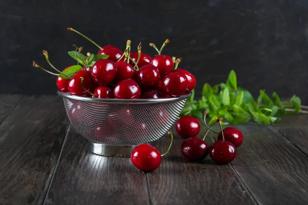 Fresh cherry in a metal sieve herbs on dark wooden background. — Stock Photo, Image