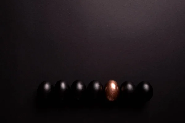 One golden egg and black eggs lie in a row on black background with copy space