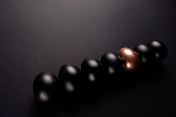 One golden egg and black eggs lie in a row on black background with copy space