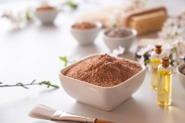 Kırmızı kil tozlu seramik kase, ev yapımı yüz ve vücut maskesi için malzemeler ya da fırça ve beyaz arka planda çiçek açan kiraz filizleri. Spa ve vücut bakımı kavramı.