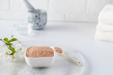 Kırmızı kil tozlu seramik kase, ev yapımı yüz ve vücut maskesi için malzemeler ya da fırça ve beyaz arka planda çiçek açan kiraz filizleri. Spa ve vücut bakımı kavramı.