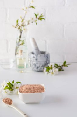 Kırmızı kil tozlu seramik kase, ev yapımı yüz ve vücut maskesi için malzemeler ya da fırça ve beyaz arka planda çiçek açan kiraz filizleri. Spa ve vücut bakımı kavramı.