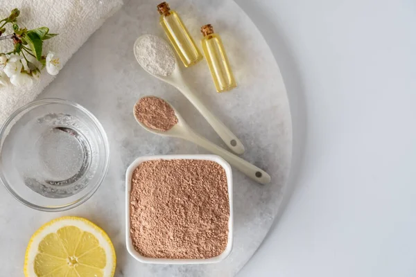 Ceramic bowl with red clay powder, ingredients for homemade facial and body mask or scrub and fresh sprig of flowering cherry on white background. Spa and bodycare concept.