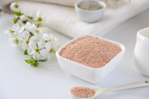 Ceramic bowl with red clay powder, ingredients for homemade facial and body mask or scrub and fresh sprig of flowering cherry on white background. Spa and bodycare concept.