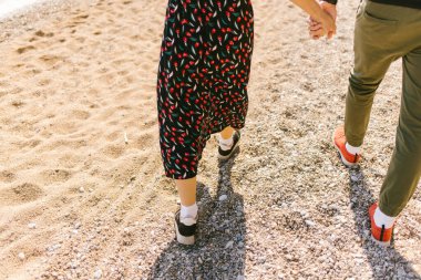 Erkek ve kız güneşli sahilde el ele yürüyorlar.