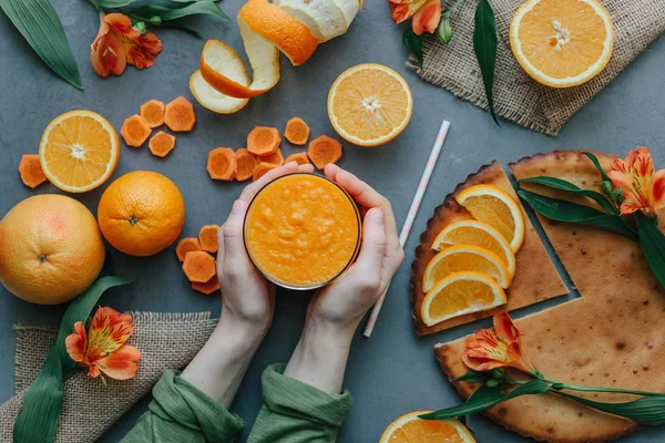 Female hands holding orange smoothie decorated with alstroemeria, halfs of orange and carrot rounds on grey table with orange pie