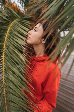 Kırmızı kazaklı kız palmiye ağacının yanında duruyor.